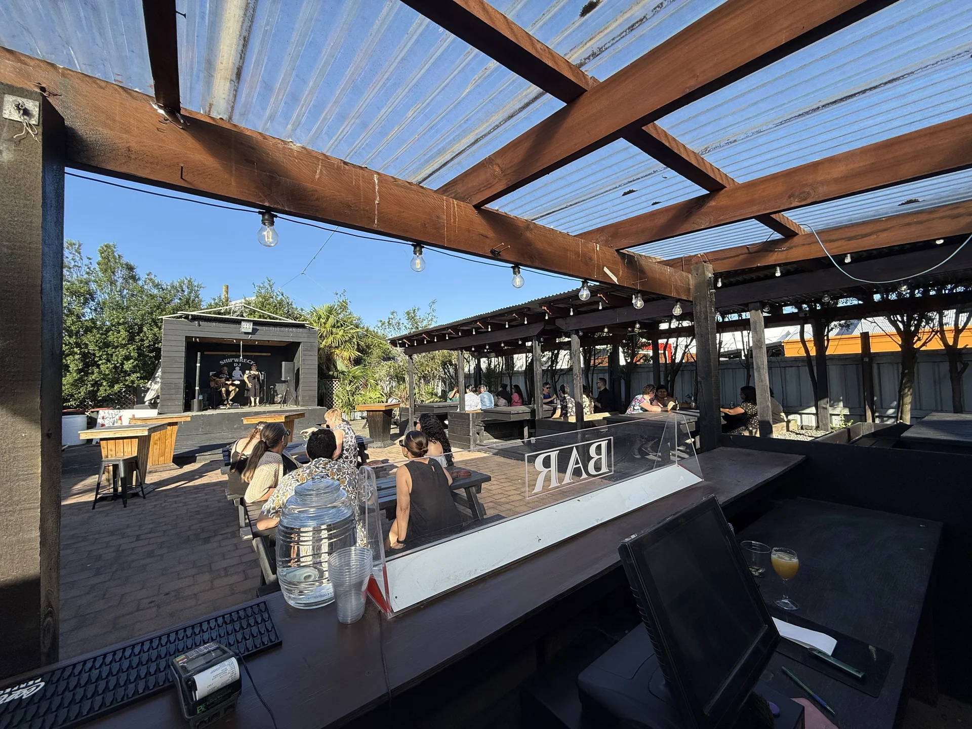 View from behind the bar across the sunny garden to the Shipwreck stage, with a duo performing and guests at tables